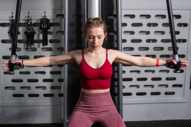 Genç atletik güzel bir kadın tam teçhizatlı bir spor salonunda göğüs makinesinde egzersiz yapıyor..