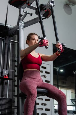 Genç atletik güzel bir kadın tam teçhizatlı bir spor salonunda göğüs makinesinde egzersiz yapıyor..