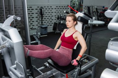 Genç, atletik bir kadın spor salonundaki bir makineye bacak presi yapıyor, bacaklarını çalıştırmak için..