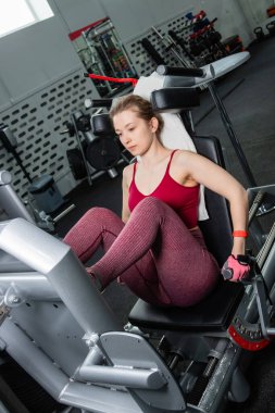 Genç, atletik bir kadın spor salonundaki bir makineye bacak presi yapıyor, bacaklarını çalıştırmak için..