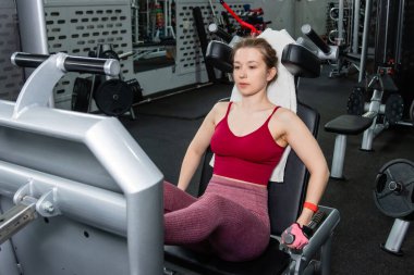 Genç, atletik bir kadın spor salonundaki bir makineye bacak presi yapıyor, bacaklarını çalıştırmak için..