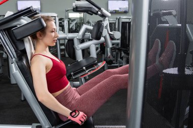 Genç, atletik bir kadın spor salonundaki bir makineye bacak presi yapıyor, bacaklarını çalıştırmak için..