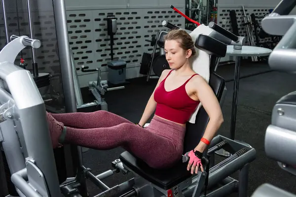 Genç, atletik bir kadın spor salonundaki bir makineye bacak presi yapıyor, bacaklarını çalıştırmak için..