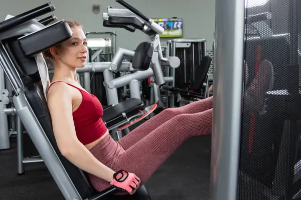 Genç, atletik bir kadın spor salonundaki bir makineye bacak presi yapıyor, bacaklarını çalıştırmak için..