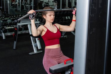 Spor salonundaki bir çekme makinesinde egzersiz yapan atletik genç bir kadın. Vücut ve kol egzersizi.