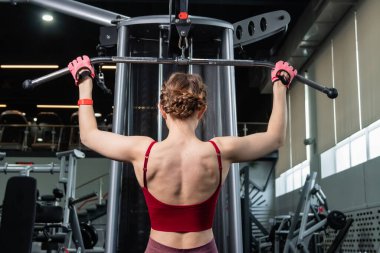 Spor salonundaki bir çekme makinesinde egzersiz yapan atletik genç bir kadın. Vücut ve kol egzersizi.