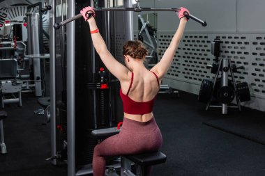 Spor salonundaki bir çekme makinesinde egzersiz yapan atletik genç bir kadın. Vücut ve kol egzersizi.