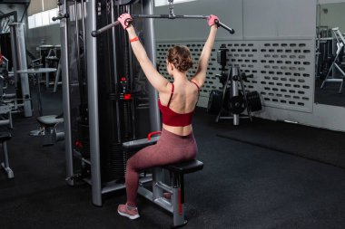 Spor salonundaki bir çekme makinesinde egzersiz yapan atletik genç bir kadın. Vücut ve kol egzersizi.