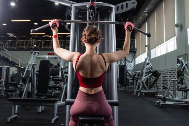 Spor salonundaki bir çekme makinesinde egzersiz yapan atletik genç bir kadın. Vücut ve kol egzersizi.