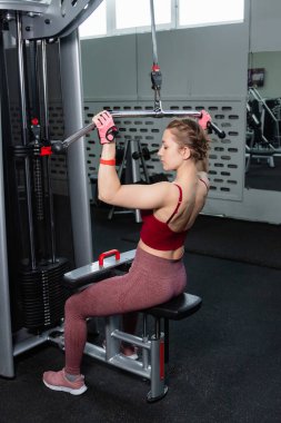 Spor salonundaki bir çekme makinesinde egzersiz yapan atletik genç bir kadın. Vücut ve kol egzersizi.