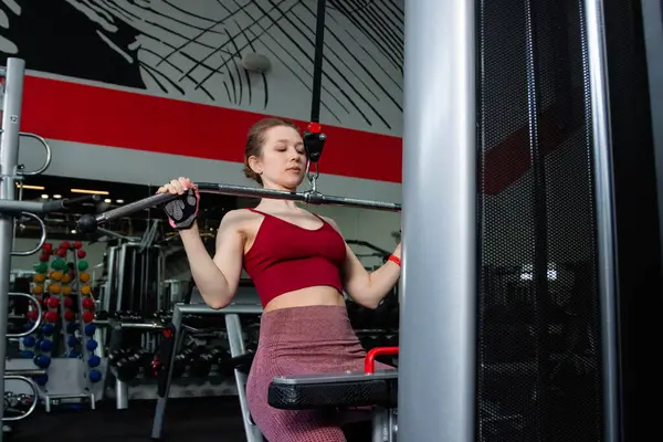 Spor salonundaki bir çekme makinesinde egzersiz yapan atletik genç bir kadın. Vücut ve kol egzersizi.