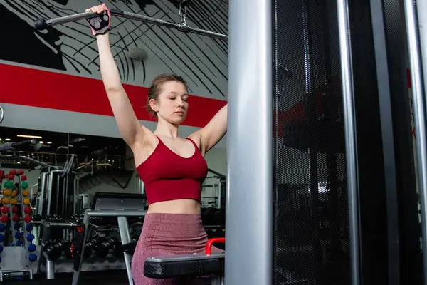 Spor salonundaki bir çekme makinesinde egzersiz yapan atletik genç bir kadın. Vücut ve kol egzersizi.