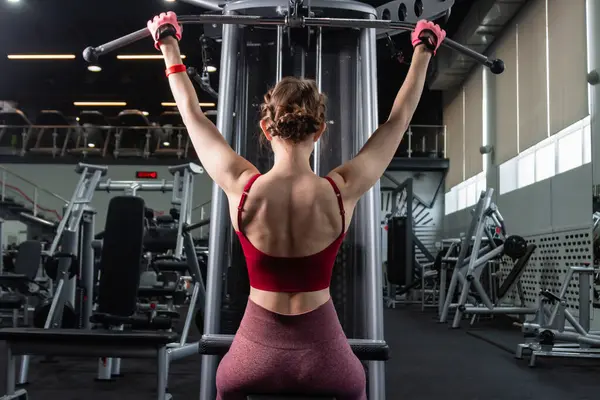 Spor salonundaki bir çekme makinesinde egzersiz yapan atletik genç bir kadın. Vücut ve kol egzersizi.