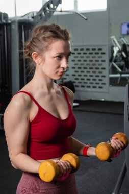 Spor salonunda arka planda egzersiz makineleri ile kol ve kolları çalıştırmak için dambıl egzersizi yapan genç bir kadın..
