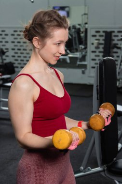 Spor salonunda arka planda egzersiz makineleri ile kol ve kolları çalıştırmak için dambıl egzersizi yapan genç bir kadın..