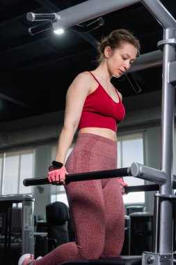 Egzersiz makinesinin yanında duran genç atletik kadın, triseps egzersizi yapıyor, spor salonunda paralel bar şınav çekiyor..