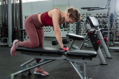 Spor salonunda halterlerle egzersiz yapan, arka planda egzersiz makineleri olan genç bir kadın..