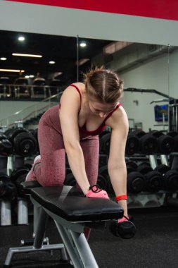 Spor salonunda halterlerle egzersiz yapan, arka planda egzersiz makineleri olan genç bir kadın..