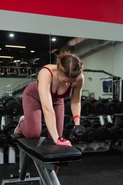 Spor salonunda halterlerle egzersiz yapan, arka planda egzersiz makineleri olan genç bir kadın..