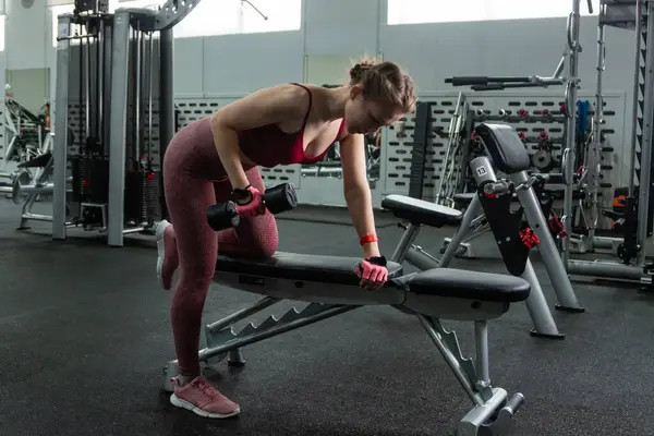 Spor salonunda halterlerle egzersiz yapan, arka planda egzersiz makineleri olan genç bir kadın..