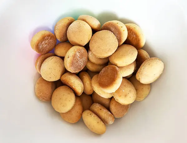 stock image A close-up view of a white bowl filled with round, golden-brown biscuits or cookies. The biscuits have a smooth texture with a slightly darker edge, indicating a well-baked crust. The bowl is placed on a white surface. 