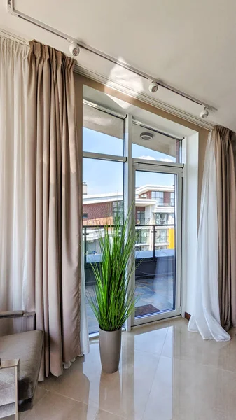 stock image Curtains on full height windows in the interior of the house. Curtain rods installed in the ceiling at an angle to the horizon. Cornices with beige drapes and white tulles. Interior details close-up. 