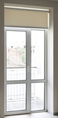 Parlak, beyaz bir balkon kapısında paten körlüğü güneşli bir şehir manzarasına açılıyor ve iç mekâna parlak ve havadar bir his veriyor. Açık güneş kremi perdesi pencereyi kapalı tutuyor..