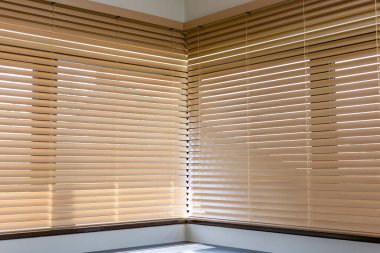 Bej ahşap panjurların doğru açıda buluştuğu köşe manzara. Işık hafifçe motorlu tahta perdelerin arasından süzülür, pencerenin pervazına gölge düşürür ve sıcak iç kısımları güçlendirir..