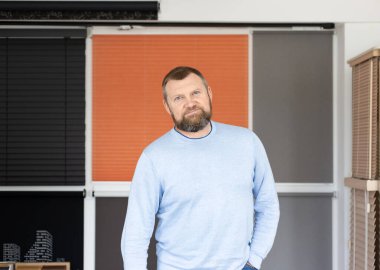 Küçük bir işletme sahibi, güneşlik sistemi galerisinde güven içinde duruyor. Paten panjurları, plili güneşlikler ve ahşap perdelerle çevrelenmiş halde, o gururla sunar kendi özel pencere kaplama çözümleri.