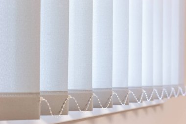 Yumuşak kumaş çatalları ve bağlı alt zincir ile dikey perdelerin yakın görüntüsü, dokuyu ve ayrıntıları gösterir. Pencere kaplaması, modern, minimalist bir iç mekanda ışık kontrolü ve gizlilik sağlar..