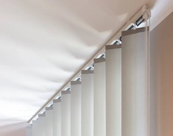 Bej dikey jaluzilerin üst kısmı ayrıntılı olarak gösteriliyor, demiryolu ve kontrol zinciri mekanizmasını vurguluyor, modern ev veya ofis iç mekanları için hem güneş koruması hem de gizlilik sağlıyor.