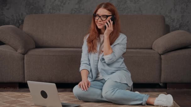 赤い髪の女性が携帯電話を持ってる友人と話してるアパートのリビングルームの床に座ってる — ストック動画