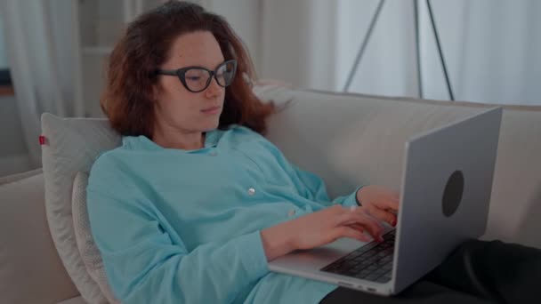 一个女人坐在沙发上 忙于笔记本电脑 她专注于屏幕 打字和滚动通过内容 布景是一个舒适的客厅 免版税图库视频