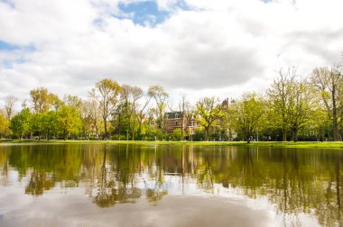 Sakin bir göl, Amsterdam 'ın parkındaki ağaçları ve bir binayı yansıtır. Doğanın güzelliği, hareketli şehirlerde bile huzurlu bir kaçış sağlar..