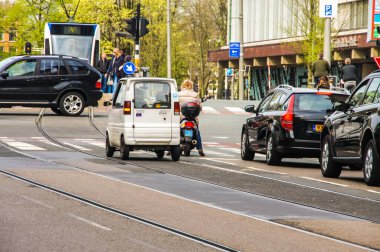 A humorous scene in Amsterdam traffic. A woman on a motorcycle follows a very small car, highlighting the size difference in a lighthearted way. clipart