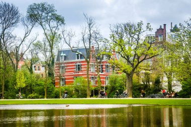 Amsterdam 'da manzaralı bir bisiklet turu. Bisikletçiler, arka planda kırmızı bir bina bulunan gölün yanındaki patikada gezinerek doğada boş vakit geçirmekten hoşlanırlar..