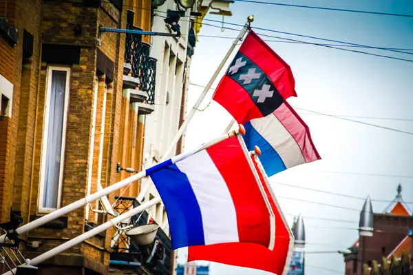 stock image Two colorful flags flutter on a brick building lining a street in Amsterdam. A vibrant display of national pride and international relations, symbolizing the city's openness to the world.