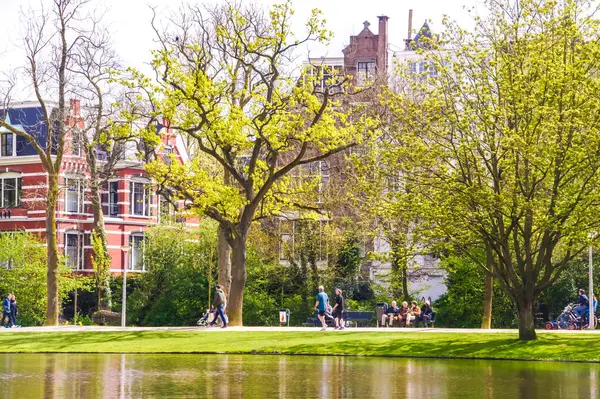 stock image A scenic park in Amsterdam. People in casual clothes stroll by a pond, their smiles reflecting the beauty of the surroundings. A leisurely escape in nature, or perhaps a group exercise session.