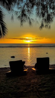 Beach chairs face a vibrant sunset over the ocean in the Cook Islands. Calm waves reflect the golden light. Tranquil moment of relaxation by the water. Escape from everyday life. clipart