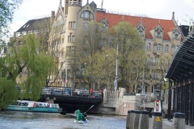 Mavi gömlekli ve kot pantolonlu bir adam Amsterdam 'da sakin, mavi bir kanalda kano kullanıyor. Modern yüksek katlı bir binanın sakin bir su yolu ile yan yana gelmesi hem huzurlu hem de huzur verici bir sahne yaratır.