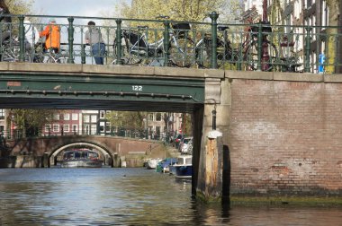 Amsterdam 'daki turistler, kenarı 122 numaralı yeşil köprüyü doldurdular. Köprü, kayıklarla benekli canlı mavi bir kanala bakıyor. Bisikletler köprünün korkuluklarına yaslanıyor.,
