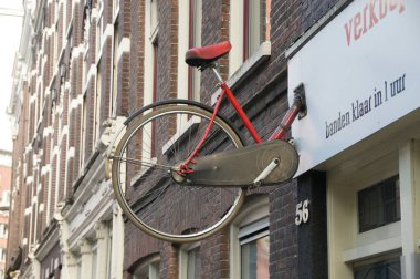 Amsterdam 'da parlak kırmızı bir bisiklet yıpranmış tuğla bir duvarda baş aşağı asılı duruyor. Bisiklet reklamlarının altına yazılan kalın Hollandaca metin bisiklet tamir servisleri, yakındaki bir bisiklet dükkanını işaret ediyor..