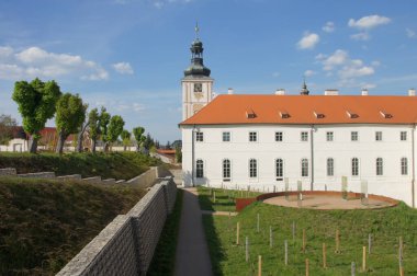 Cizvit Koleji, Kutna Hora. Kırmızı çatısı ve yüksek kulesi olan beyaz bir bina. Ağaçları ve çalıları olan taş duvar. Ağaçlarla çevrili bir tümsek. Çek Cumhuriyeti. Trajik olayların anısına. Aynı zamanda...