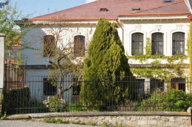 Fotoğraf beyaz yüzlü ve kırmızı çatılı bir binayı gösteriyor. Binanın önünde bir ağaç ve bazı çalılar var. Mülkü çevreleyen özel bir çit var. Bina sessiz bir yerde.