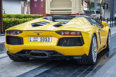 Işıldayan sarı bir Lamborghini Aventador, Dubai plakası güneşte parlıyor, bir otobüsün önüne park edilmiş. Bu sahne, müsrif Dubai şehrinin zenginliğini haykırıyor. Görüntü...
