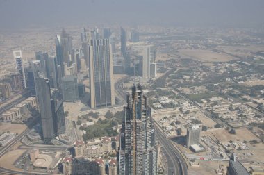 Dubai 'nin panoramik bir görüntüsü altınızda beliriyor, bir gökdelenin baş döndürücü yüksekliğinden çekiliyor. Güneş ışığı şehrin modern siluetini tanımlayan sonsuz cam ve çelik cephelerde parlıyor..