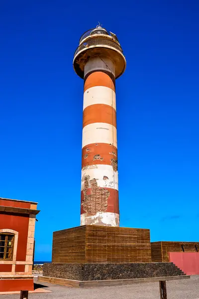 A tall lighthouse with a white stripe on it. The sky is blue and clear. The lighthouse is in the foreground and the building behind it is red