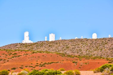 A hillside with a bunch of telescopes on it. The hill is covered in red dirt and the sky is clear and blue clipart