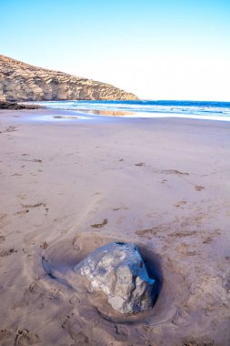 A large rock sits on the beach, surrounded by sand. The sky is clear and the water is calm. The scene is peaceful and serene, with the rock adding a sense of stability to the otherwise soft clipart