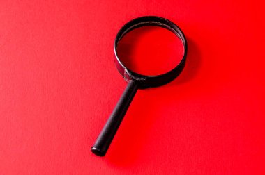 A black magnifying glass is on a red background. Concept of curiosity and exploration, as the magnifying glass is often used to examine and discover hidden details clipart